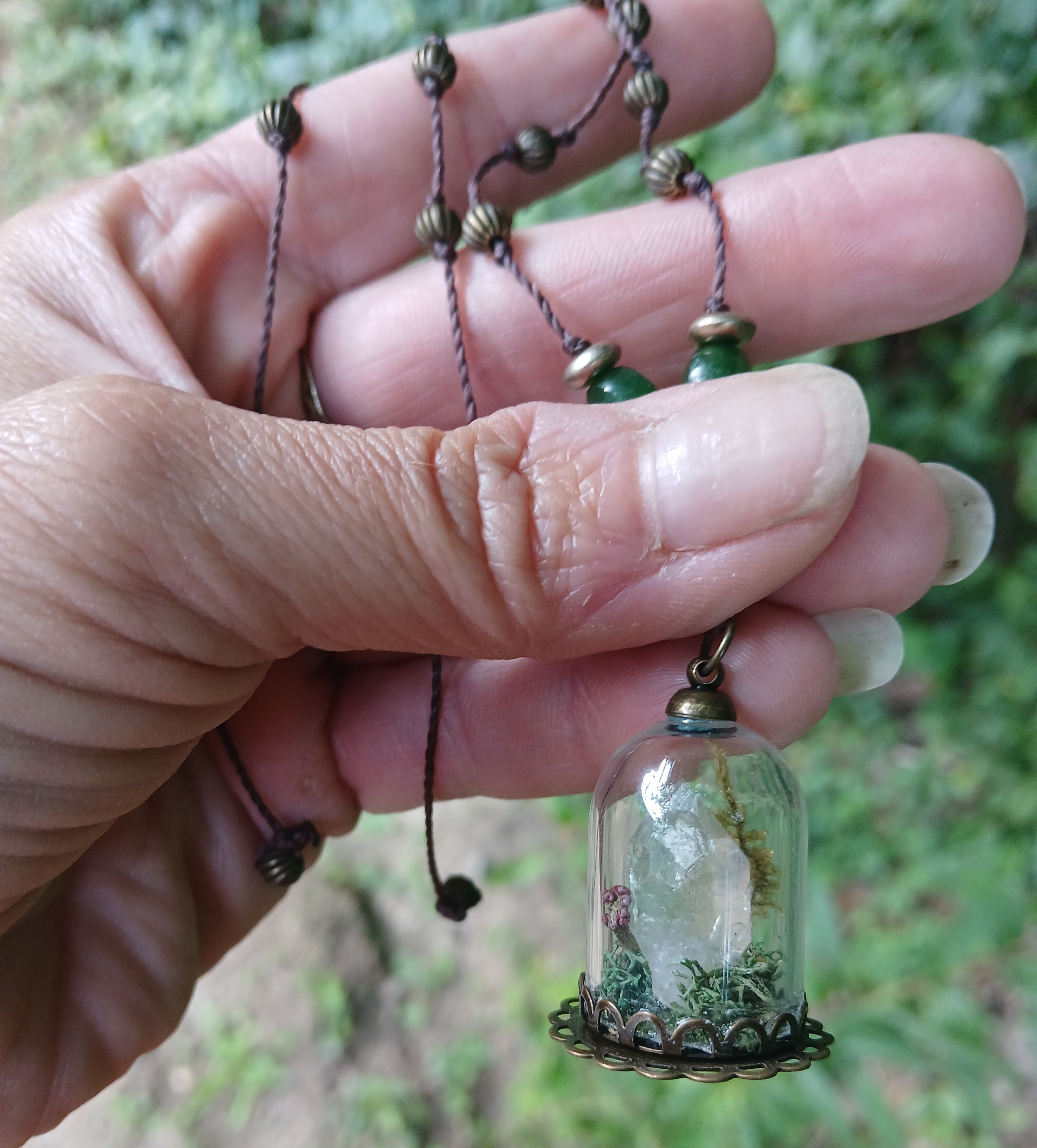 Glass cloche pendant holding a quartz point in moss, on knotted cord with green jade and brass beads